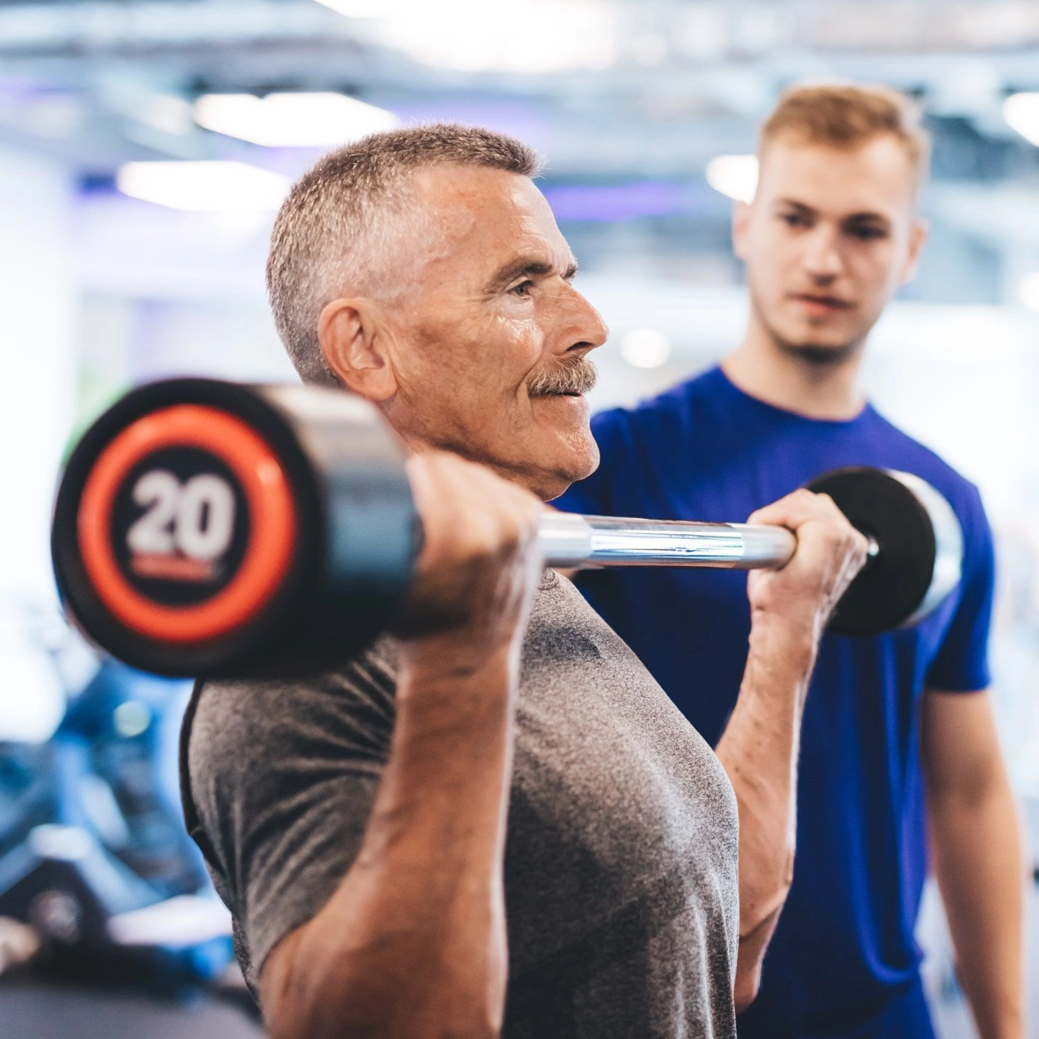 Atrium Sports e.V. - Kampfsport und Fitness – ein älterer Mann, der mit einem Personal Trainer in einem Fitnessstudio Langhantelcurls ausführt. Krafttraining bietet älteren Erwachsenen erhebliche gesundheitliche Vorteile, darunter eine verbesserte Knochendichte, ein besseres Gleichgewicht und ein gesteigertes geistiges Wohlbefinden, was dazu beiträgt, die Unabhängigkeit im Alter zu bewahren.
